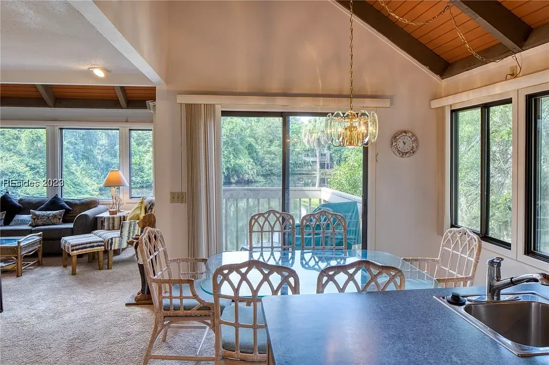 An open dining and living area with a glass-top table, wicker chairs, and vaulted wood-paneled ceiling overlooks a lagoon through large windows and a sliding glass door to a balcony.