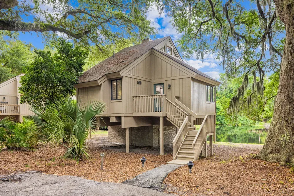 An elevated wood-sided cottage with a steep gabled roof stands on pilings beneath oak trees draped in Spanish moss, with a staircase leading up to the front deck and a lagoon visible beyond.