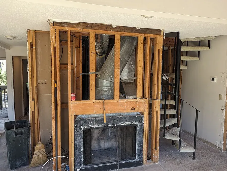 An open wall framing exposes exhaust ductwork and studs above a fireplace insert during a whole-home renovation, with a metal spiral staircase visible nearby.