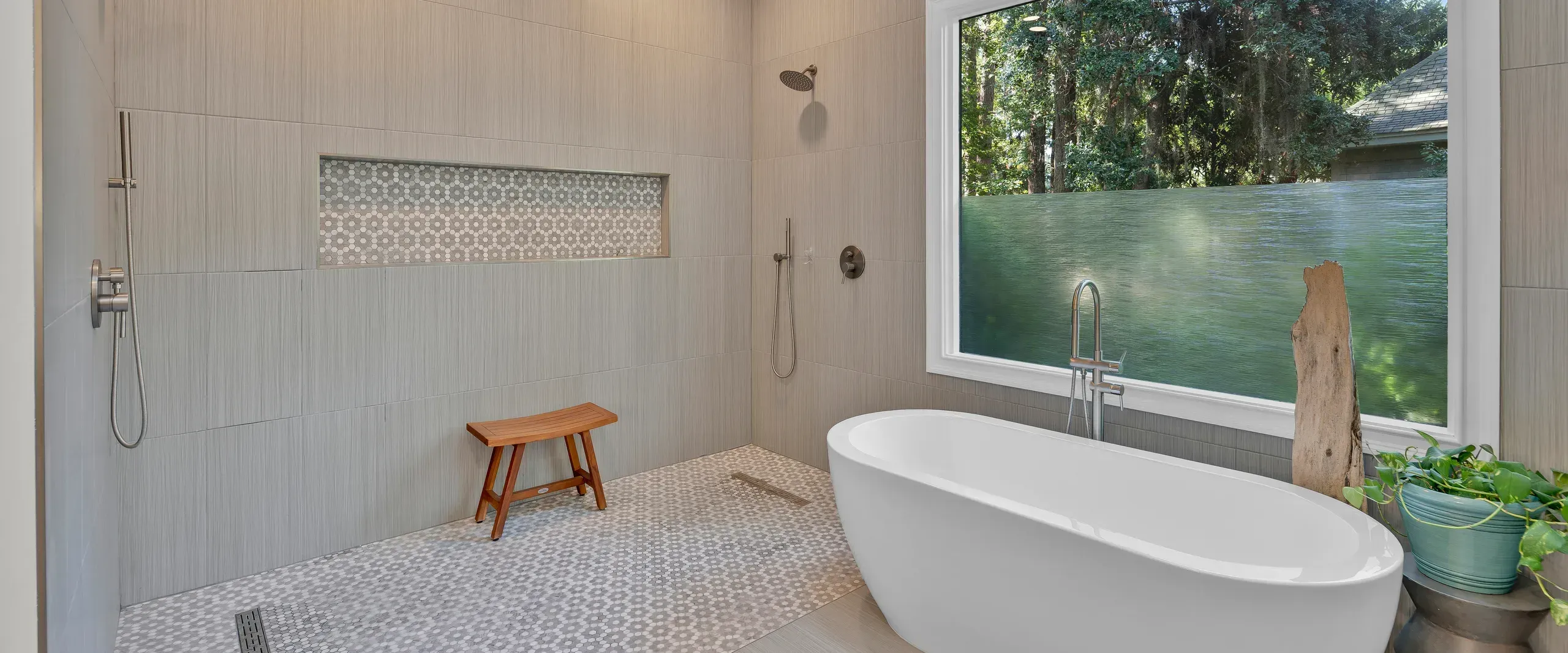 A spa-like bathroom remodel features a freestanding oval soaking tub beneath a large window overlooking wooded scenery, alongside a curbless walk-in shower with dual showerheads, a teak bench, and hexagonal mosaic tile flooring.