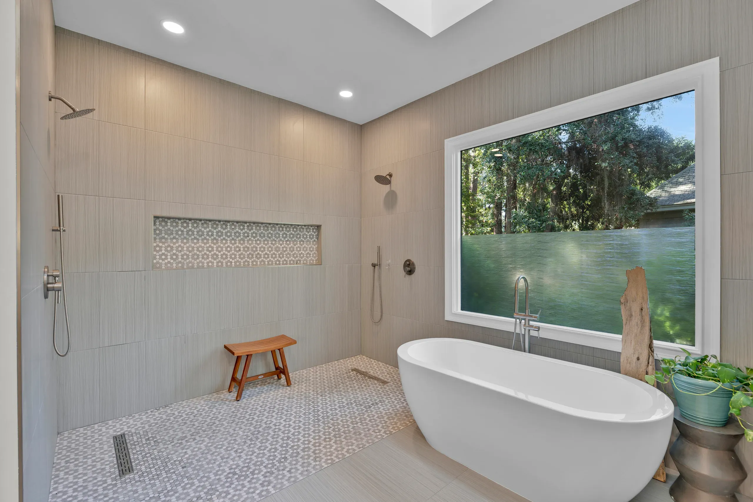 A spa-like bathroom remodel features a freestanding oval soaking tub beneath a large window overlooking wooded scenery, alongside a curbless walk-in shower with dual showerheads, a teak bench, and hexagonal mosaic tile flooring.