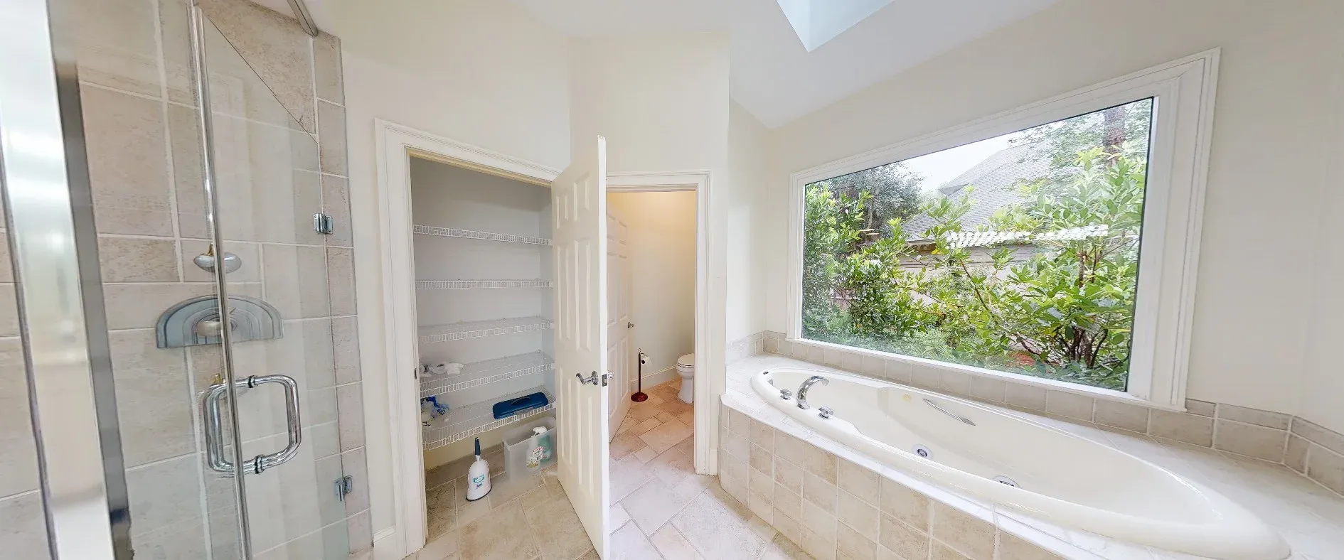 A primary bathroom features a tiled jetted soaking tub beneath a large window overlooking greenery, a glass-enclosed shower, a linen closet with wire shelving, and a separate toilet area accessed through an open door.