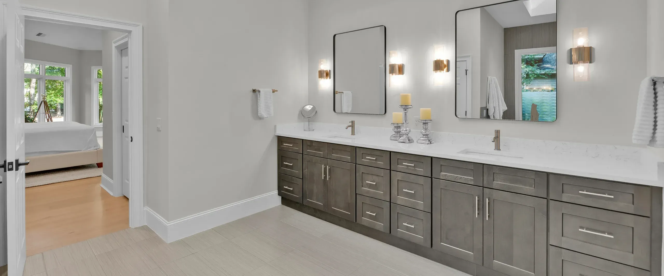 A long double-sink vanity with gray-stained cabinetry and white quartz countertops sits beneath two framed mirrors and brass sconces, with a doorway opening to a bright bedroom beyond.