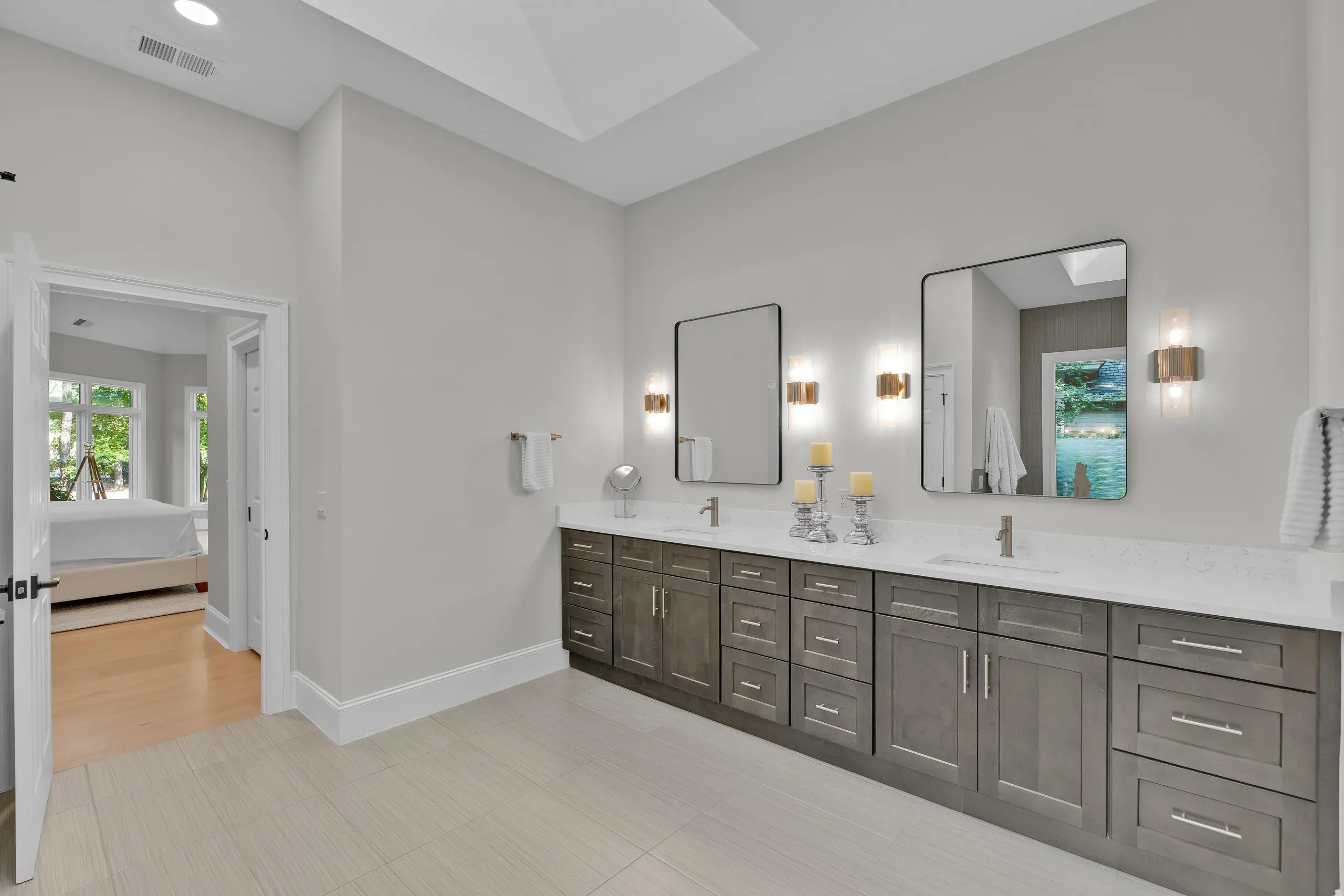 A long double-sink vanity with gray-stained cabinetry and white quartz countertops sits beneath two framed mirrors and brass sconces, with a doorway opening to a bright bedroom beyond.