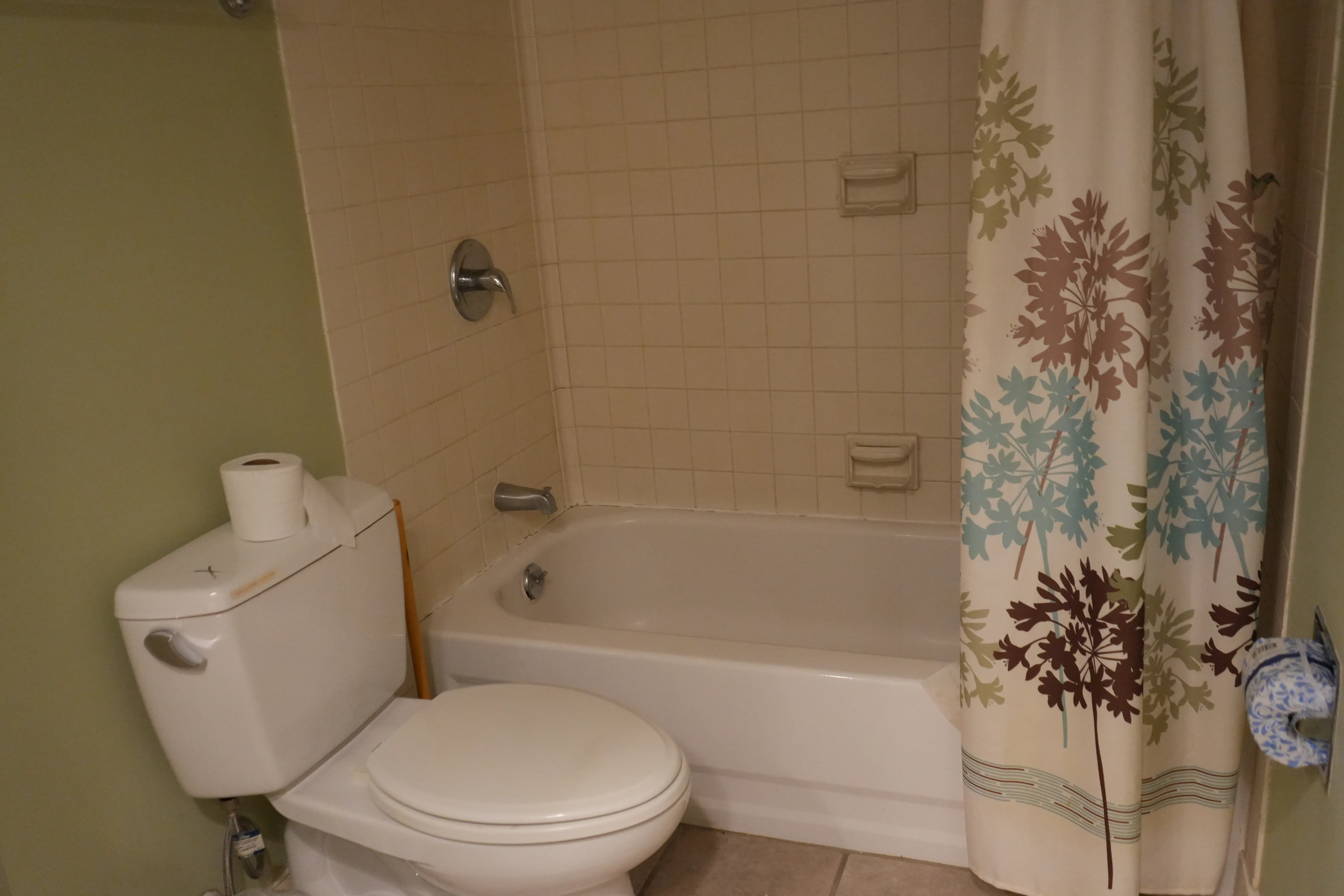 An outdated bathroom with beige tile surround, a white tub and toilet, green walls, and a floral shower curtain awaiting renovation.