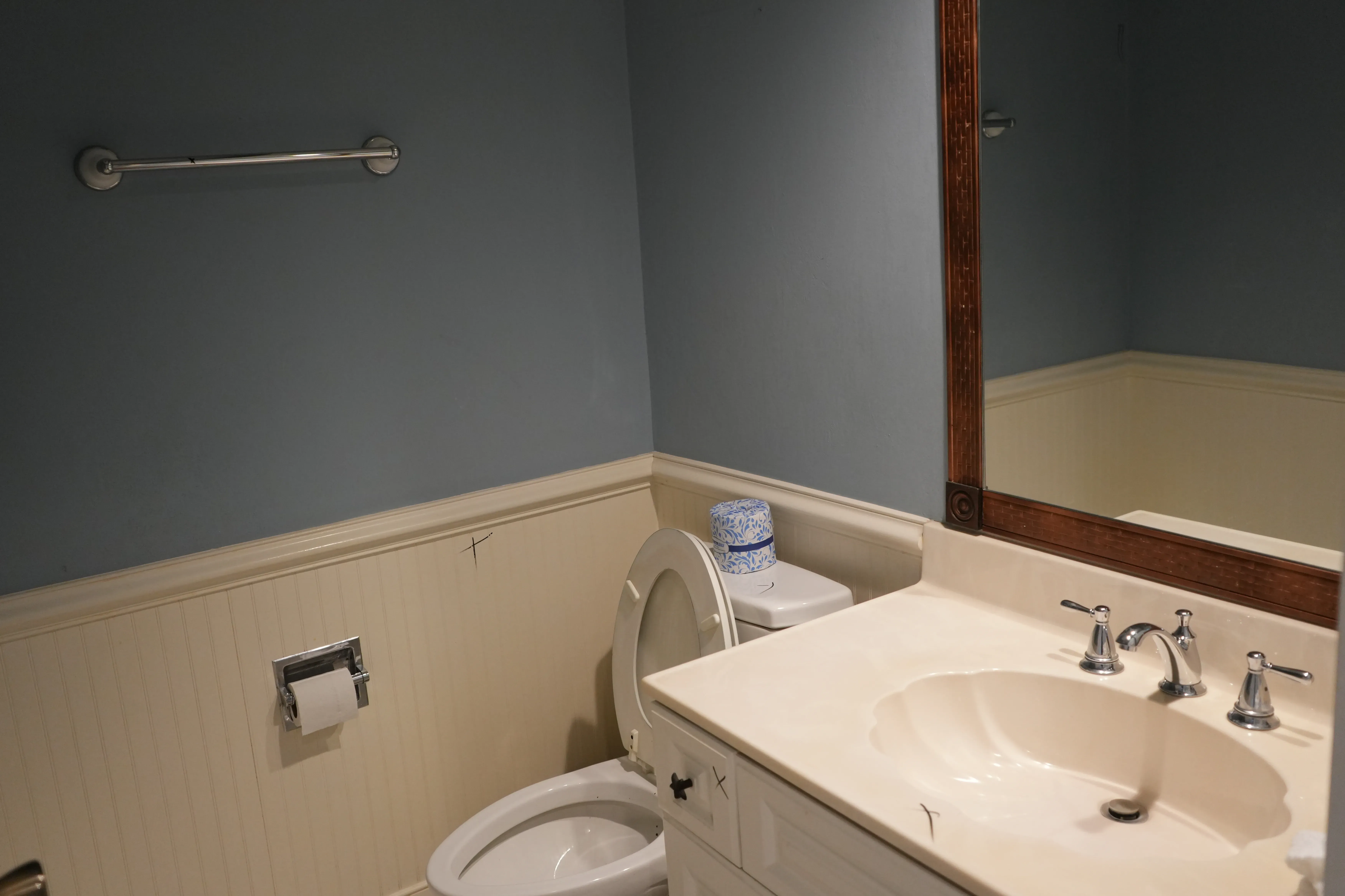 An existing bathroom shows a beadboard wainscoting, a toilet with a grab bar above it, and a beige countertop sink beneath a wood-framed mirror.