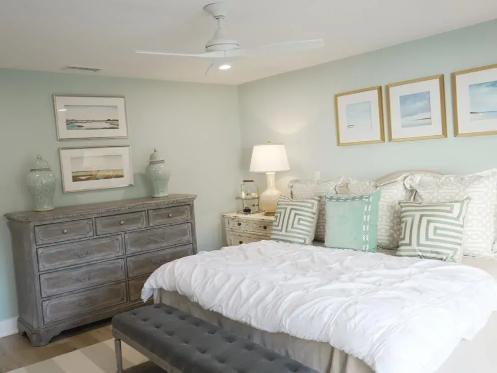 A coastal-style bedroom with soft green walls features an upholstered bed with patterned throw pillows, a weathered gray dresser topped with framed art and ceramic jars, and a white ceiling fan overhead.