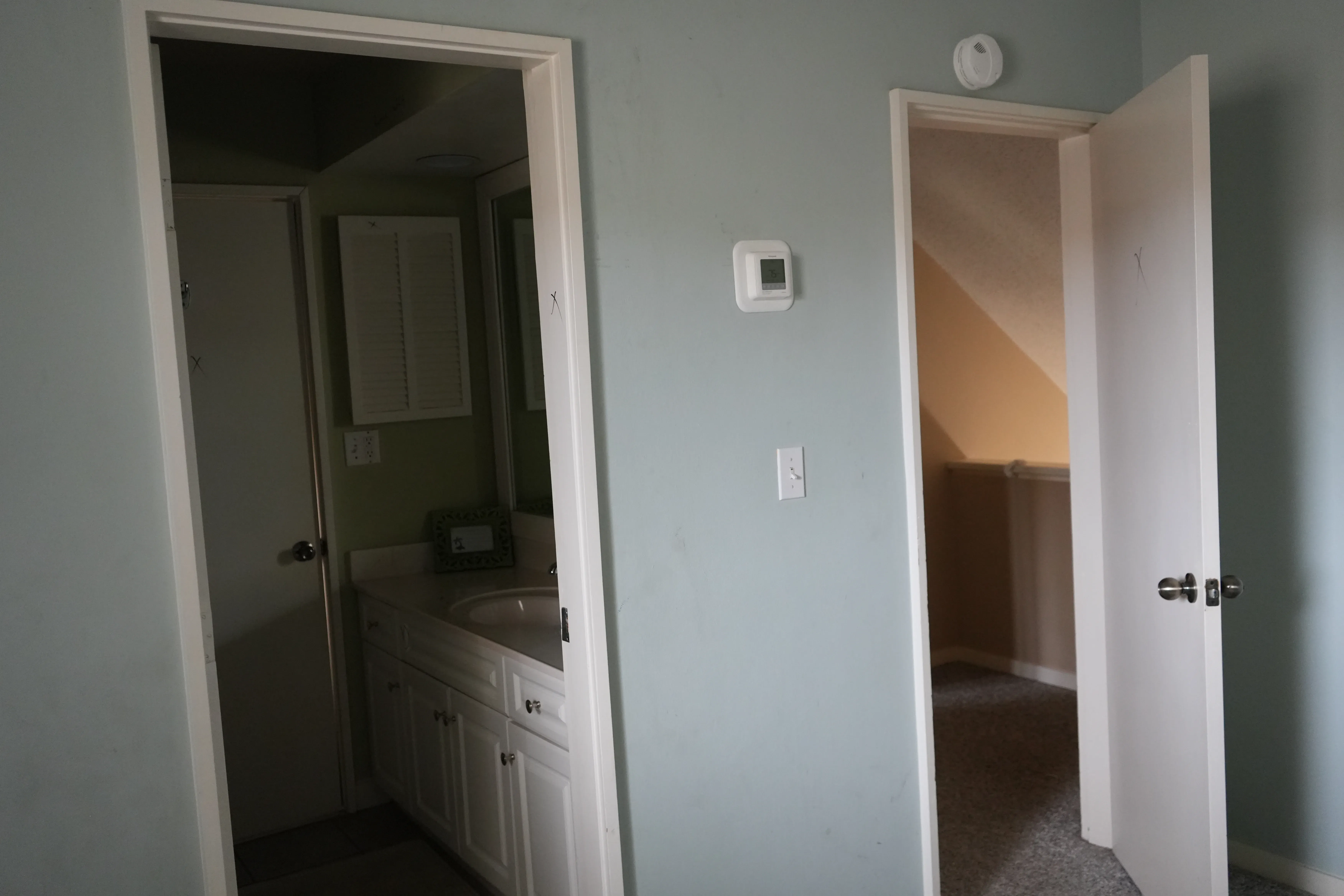 A blue-gray bedroom before renovation shows an open doorway to a small bathroom with a white vanity on one side and an open door leading to a sloped-ceiling attic space on the other.