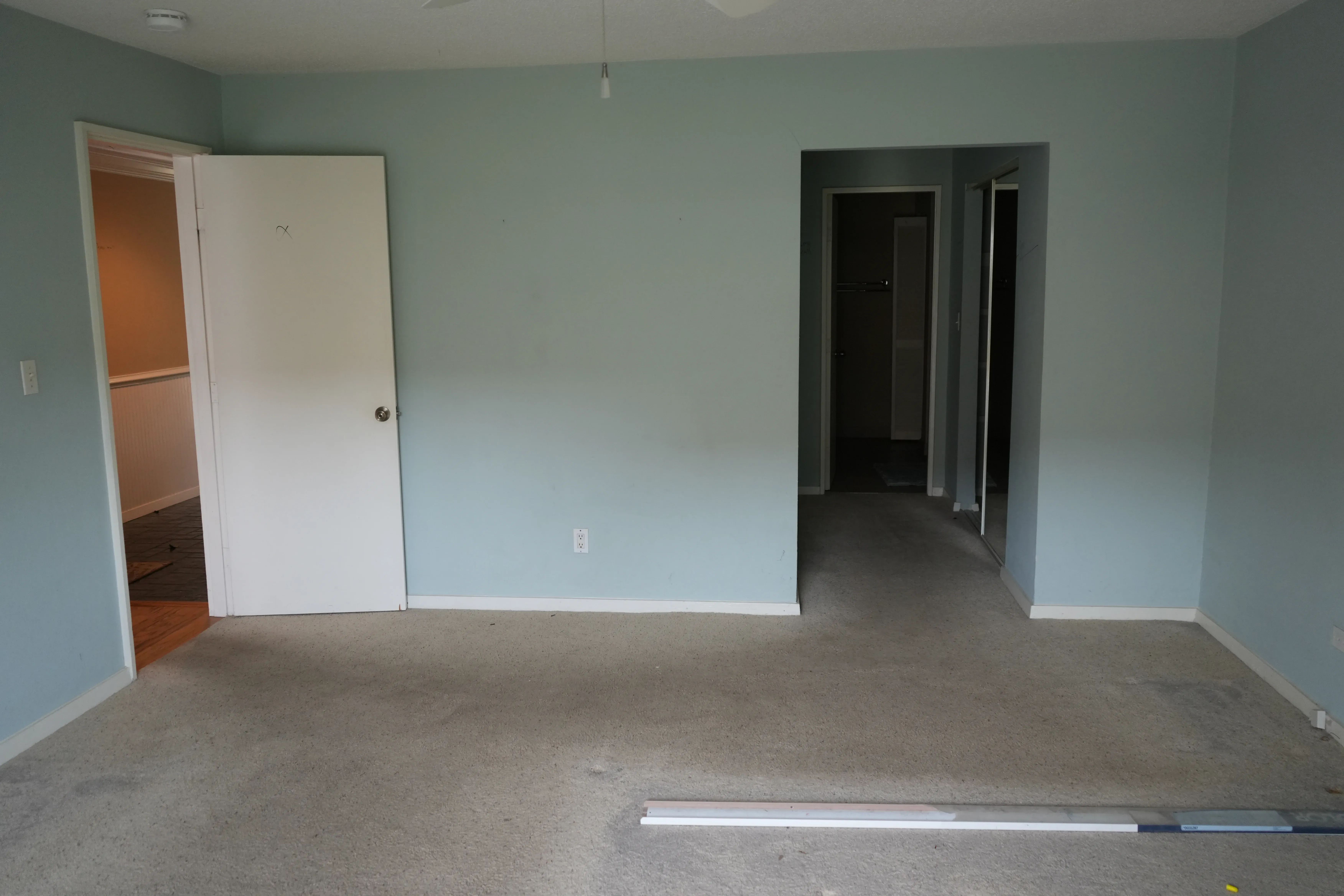 An empty bedroom with pale blue walls and gray carpeting shows an open closet door, a doorway leading to a bathroom, and trim materials staged on the floor before renovation.