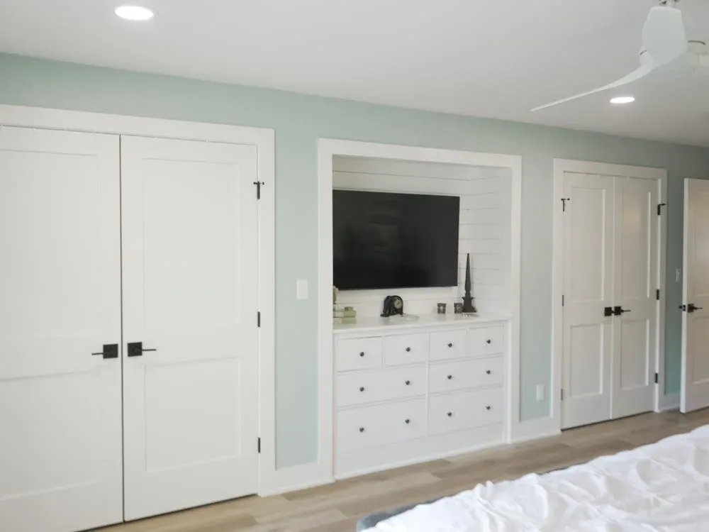 A bedroom features a built-in white dresser with a shiplap accent wall and mounted television, flanked by double closet doors and soft green walls.
