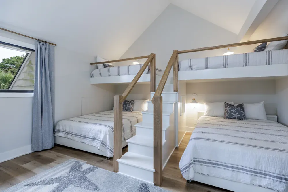 A vaulted attic bedroom features two sets of built-in bunk beds with white wood frames, wood-toned handrails, and a central staircase leading to the upper bunks, all set above a wide-plank wood floor.