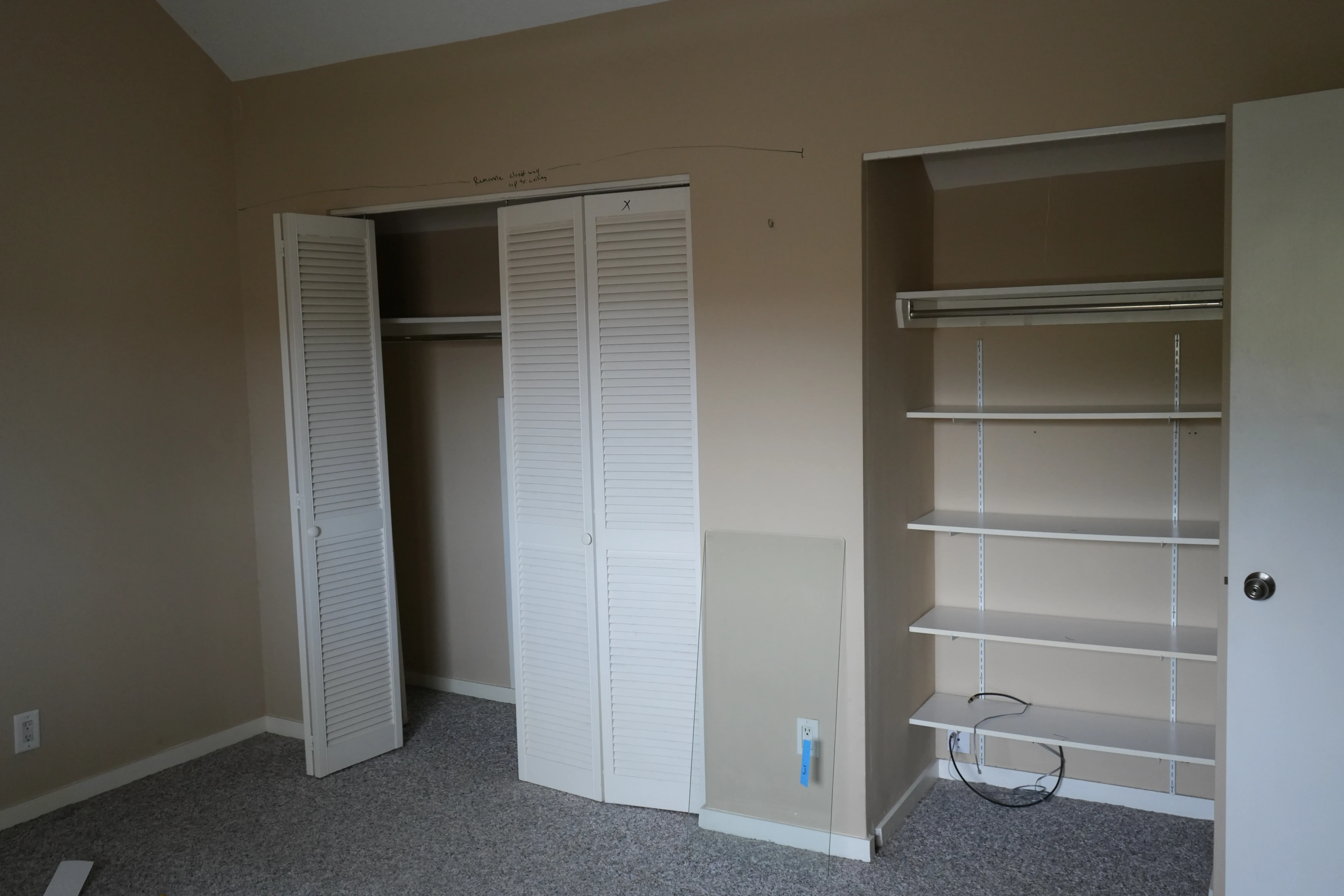 An empty bedroom with vaulted ceilings shows two closets, one with louvered bifold doors and another with open wire and laminate shelving, ready for renovation.