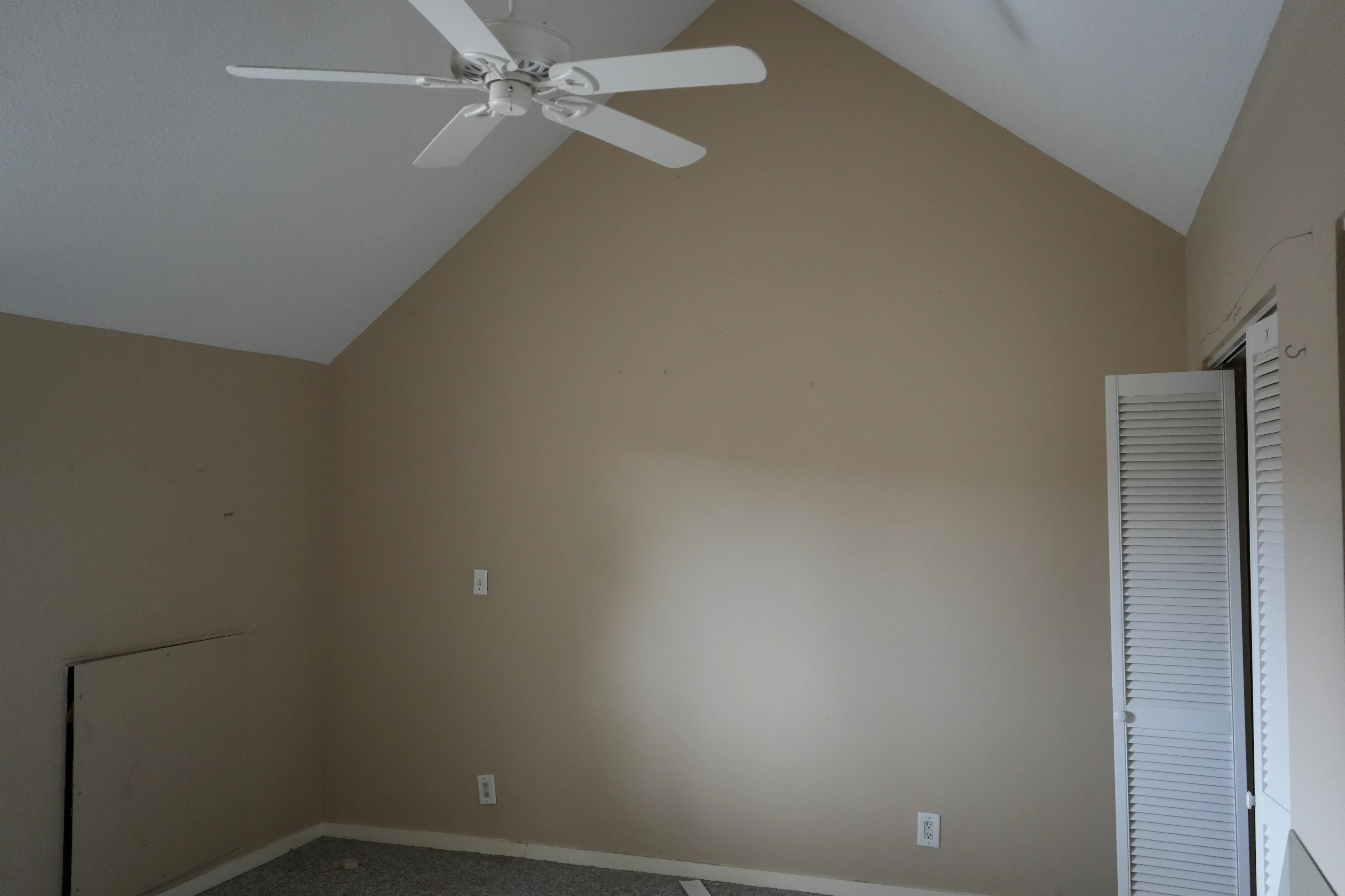 An empty bedroom under renovation shows a vaulted ceiling with a white ceiling fan, tan walls, louvered closet doors, and a partially exposed subfloor.