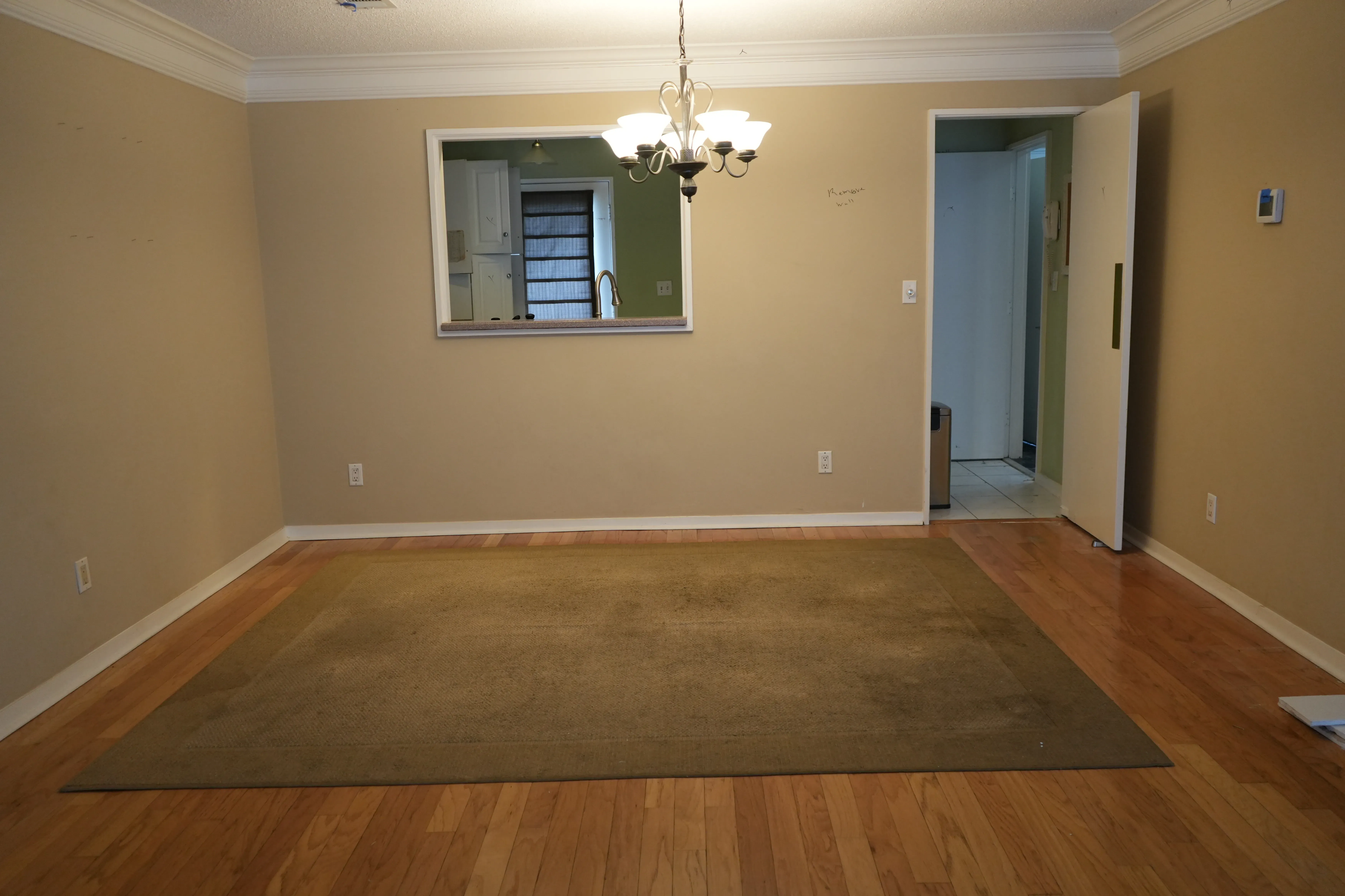 An empty dining room with hardwood floors and a green area rug features a chandelier overhead and a pass-through window opening to the kitchen beyond.