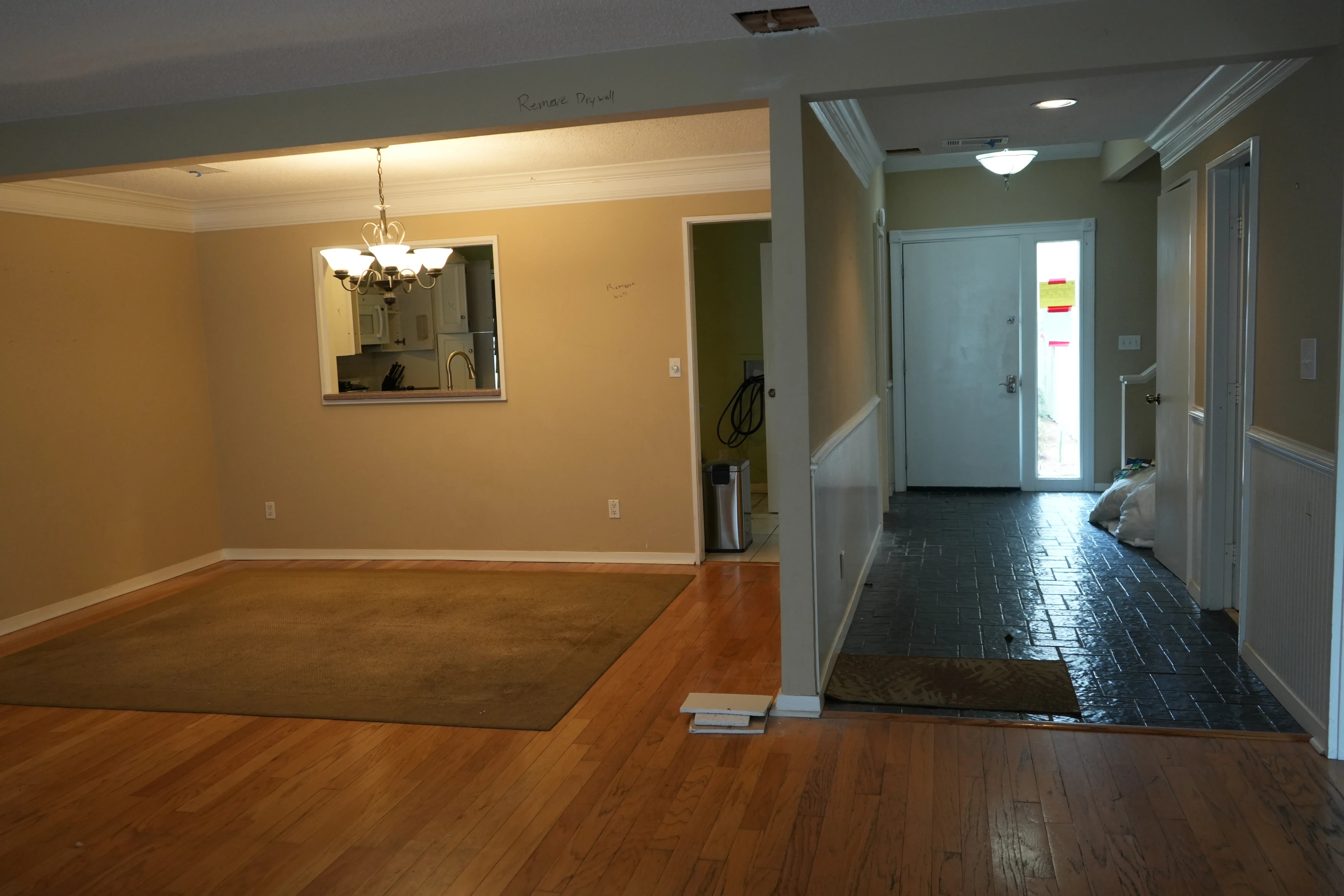 An empty dining area with hardwood floors and a chandelier opens into a tiled entryway leading to a white front door, in a home before renovation.