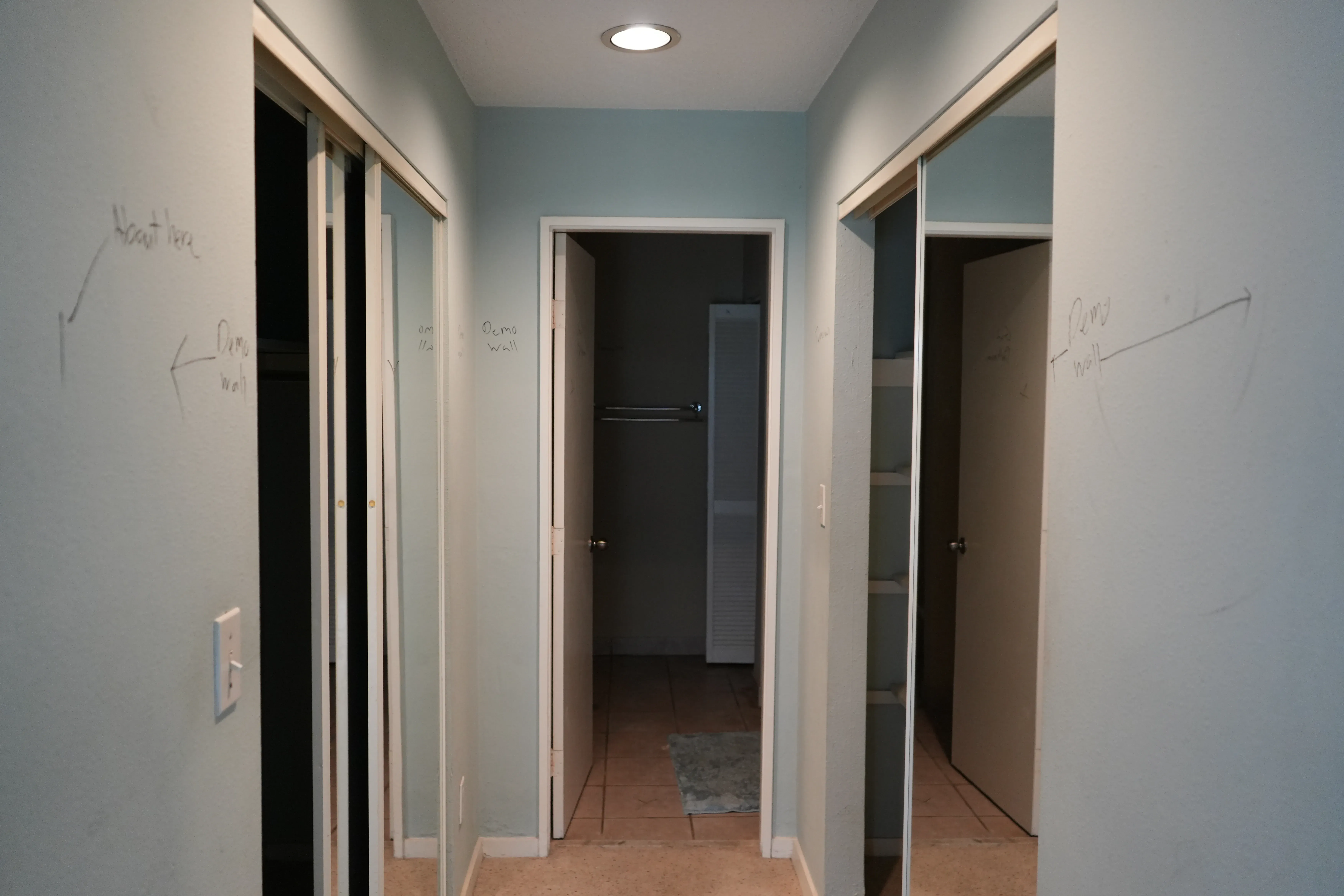 A narrow hallway with mirrored sliding closet doors on either side and pencil markings noting demolition plans on the light blue walls, leading to a doorway with a closet rod visible beyond.