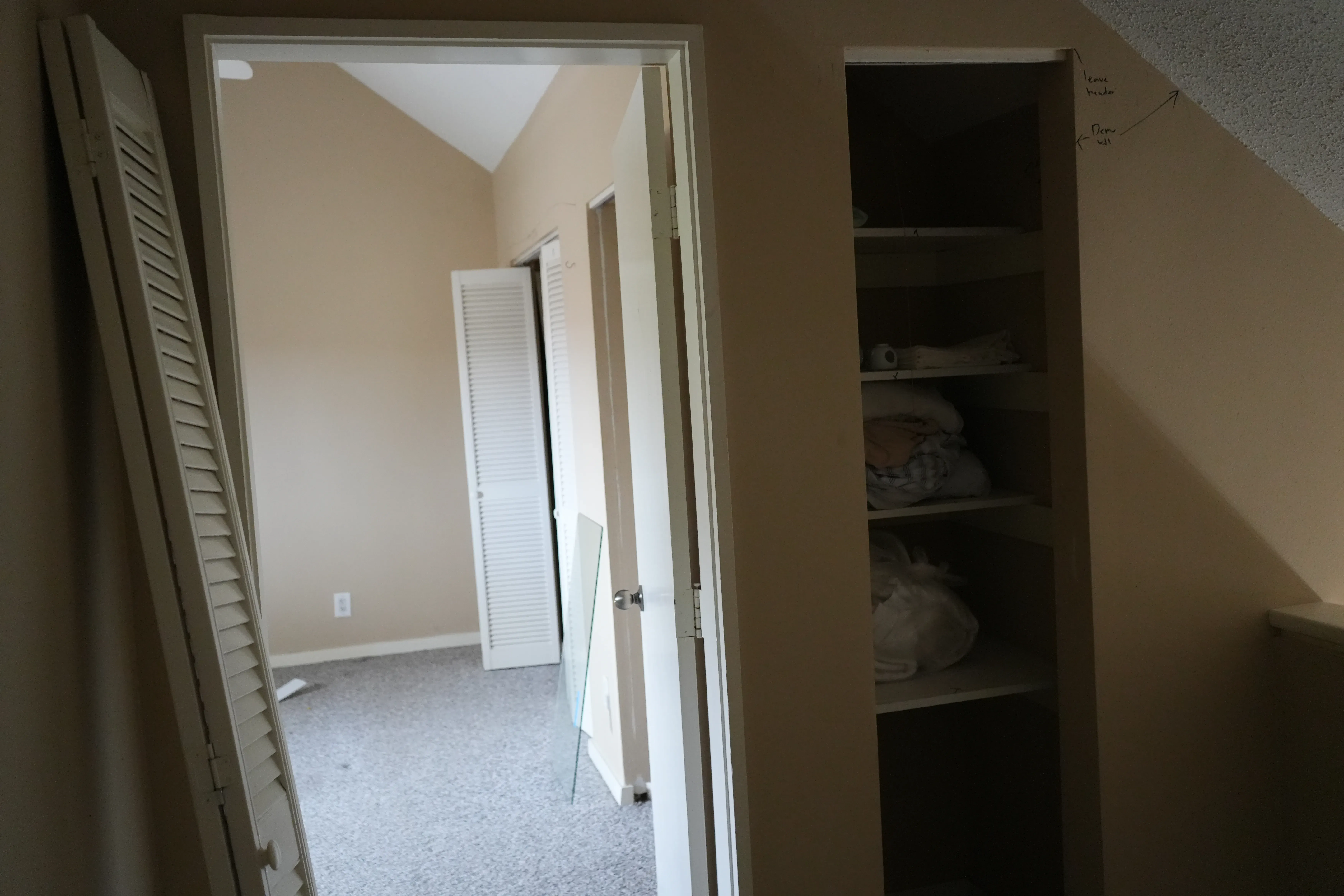 An unrenovated hallway with beige walls and gray carpet shows a linen closet with shelving beside a vaulted-ceiling bedroom with louvered closet doors, viewed past a removed shutter panel in the foreground.