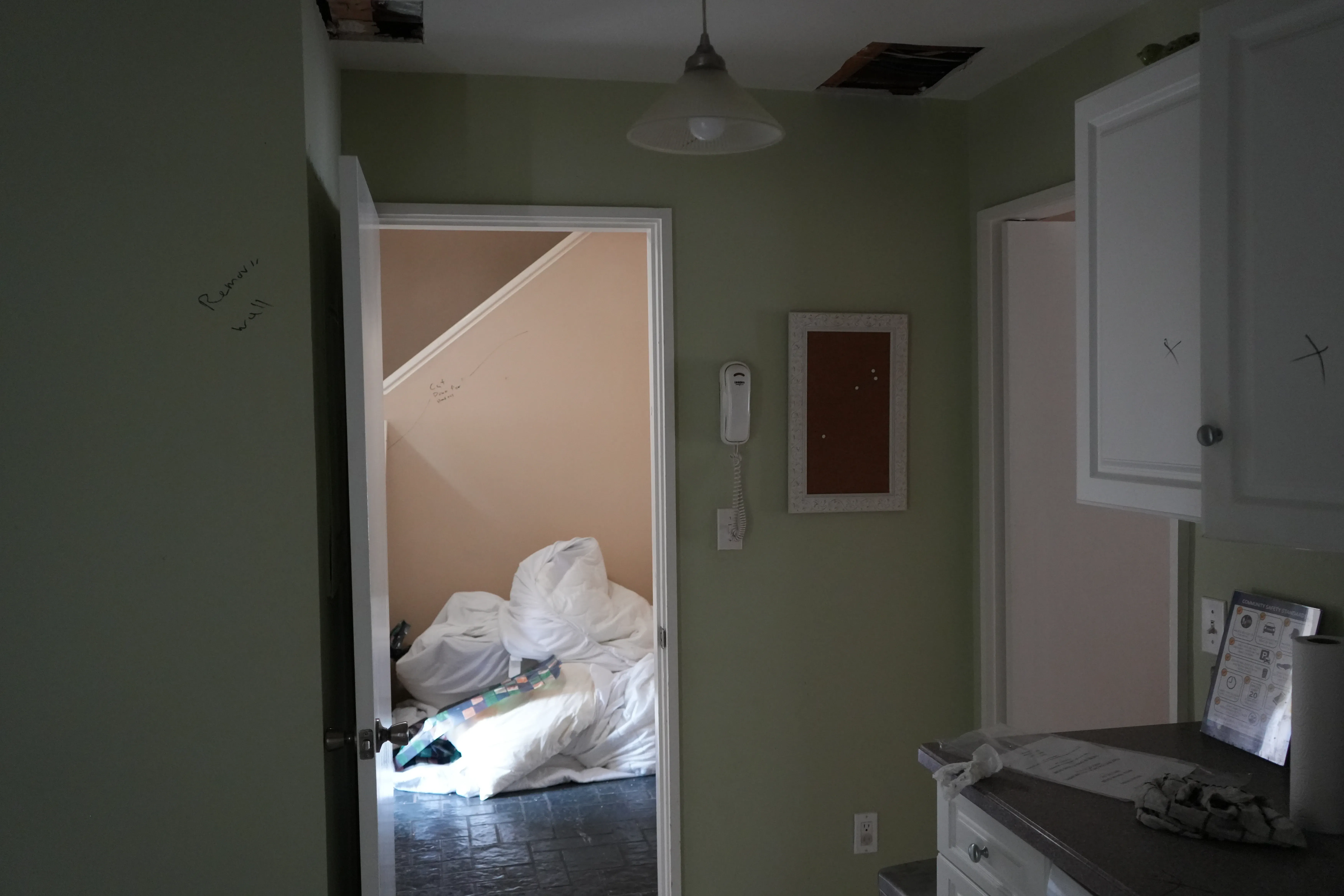 A kitchen mid-renovation shows green walls with contractor markings, an open ceiling patch, and an adjoining stairwell room filled with covered furniture and debris.