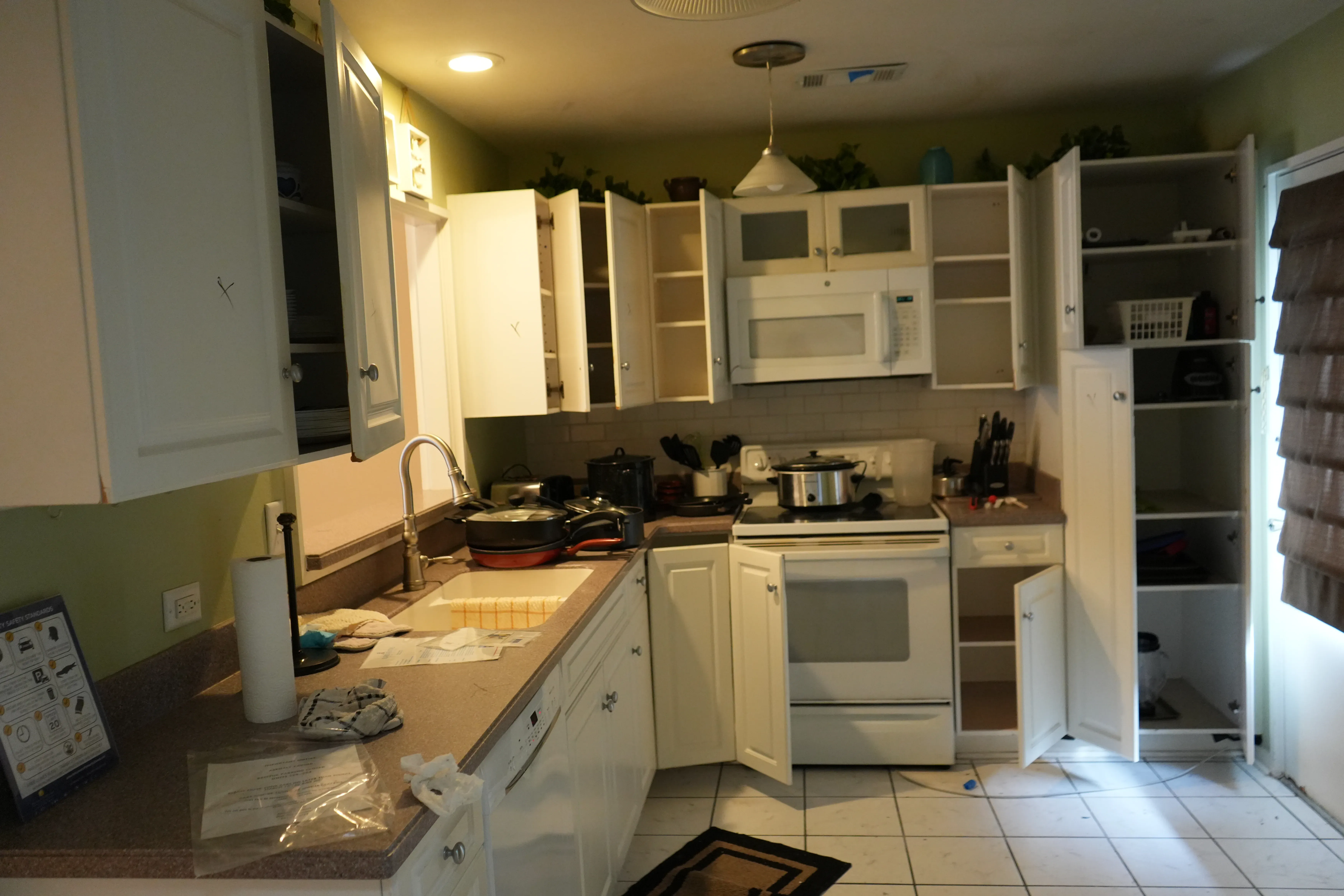 An existing kitchen with white cabinets left open shows a white range, microwave, and tan laminate countertops with a tile floor before renovation.