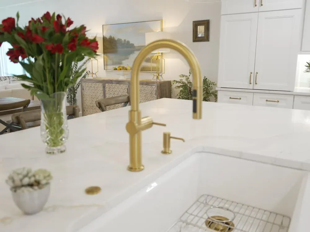 A brushed brass gooseneck faucet rises over a farmhouse sink set into a white marble kitchen island, with a vase of red roses and white cabinetry visible in the background.