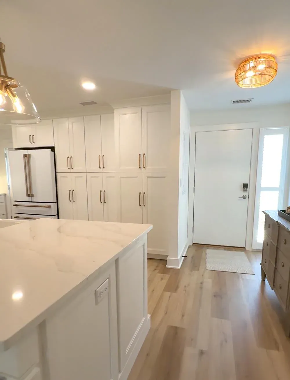 A remodeled kitchen with floor-to-ceiling white cabinetry and a marble-look island opens onto a foyer with wide-plank wood flooring and a front door flanked by a sidelight window.