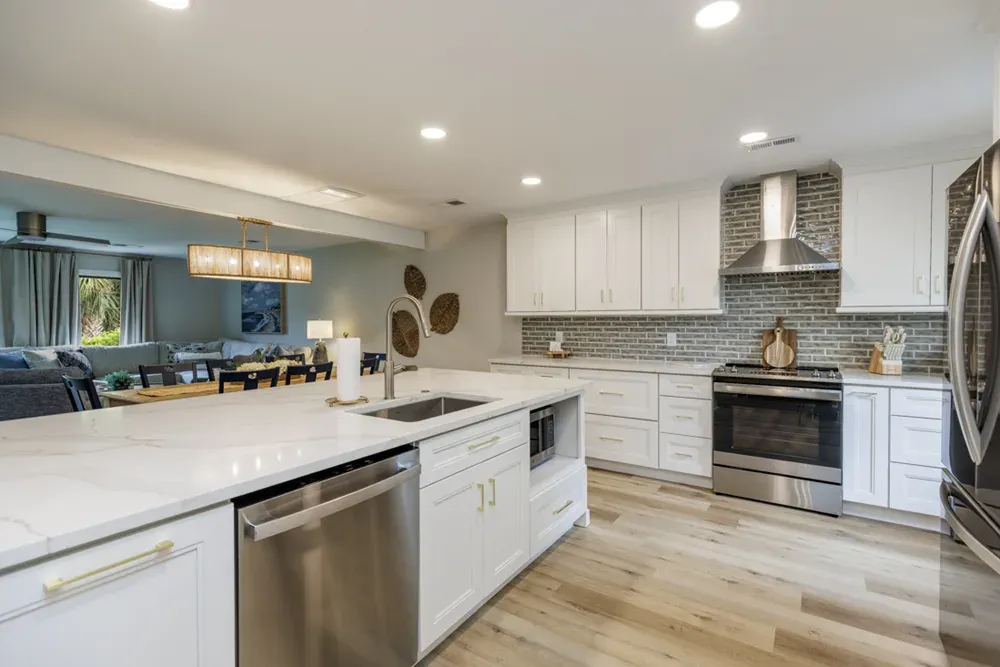 An open-concept kitchen features a large white quartz island with gold hardware, a gray brick backsplash, stainless steel appliances, and a view into an adjoining living room with a beamed ceiling.
