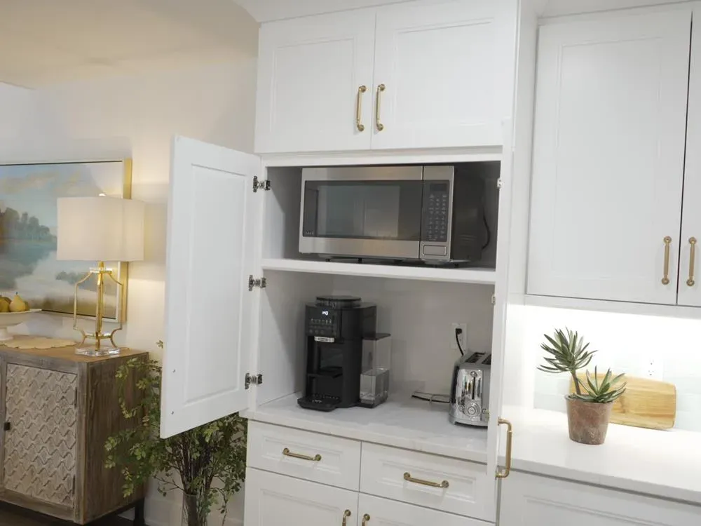 An open white cabinet in a kitchen remodel reveals a built-in microwave, coffee maker, and toaster on shelves with brass hardware and a nearby quartz counter holding a small potted succulent.