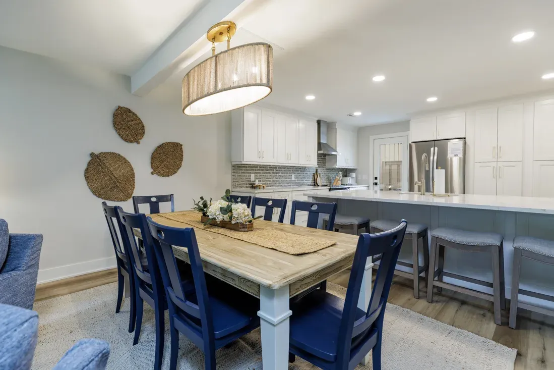 An open-concept dining area with a wooden farmhouse table and navy chairs sits beneath a drum pendant light, opening onto a white kitchen with a long quartz island and gray stone backsplash.