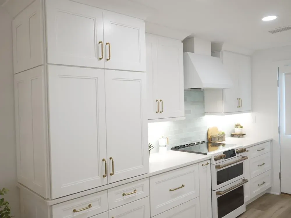 A remodeled kitchen features tall white shaker cabinets with brass pulls, a white range hood, a light green tile backsplash, and a double-oven range beside a white quartz countertop.