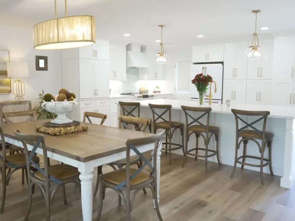 An open-concept kitchen remodel features white shaker cabinets, a long quartz-topped island with brass fixtures and cross-back barstools, and an adjoining farmhouse-style dining table beneath a woven drum pendant light.