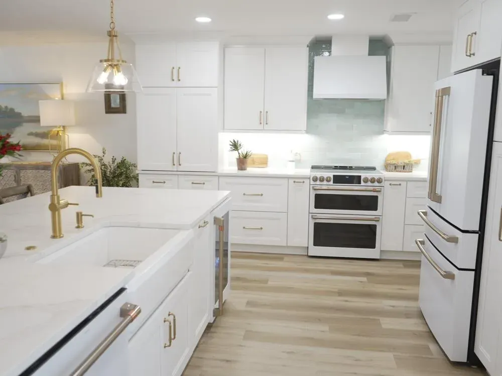 A white kitchen remodel features a large island with a farmhouse sink and gold faucet, quartz countertops, a double oven range beneath a white vent hood, and light wood flooring.