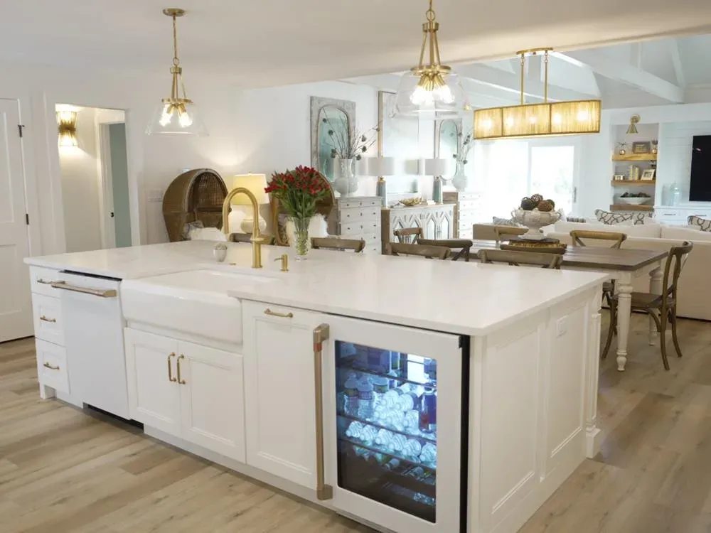 An open-concept kitchen features a large white farmhouse-sink island with brass fixtures, a built-in beverage refrigerator, and views into a dining area with vaulted ceilings.