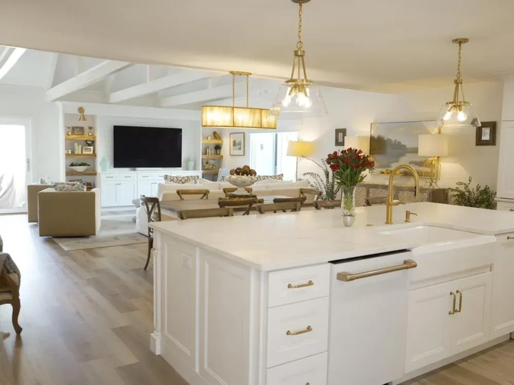 An open-concept kitchen with a white farmhouse sink and gold fixtures on a large white island opens onto a living room with vaulted white beamed ceilings and a built-in media cabinet.