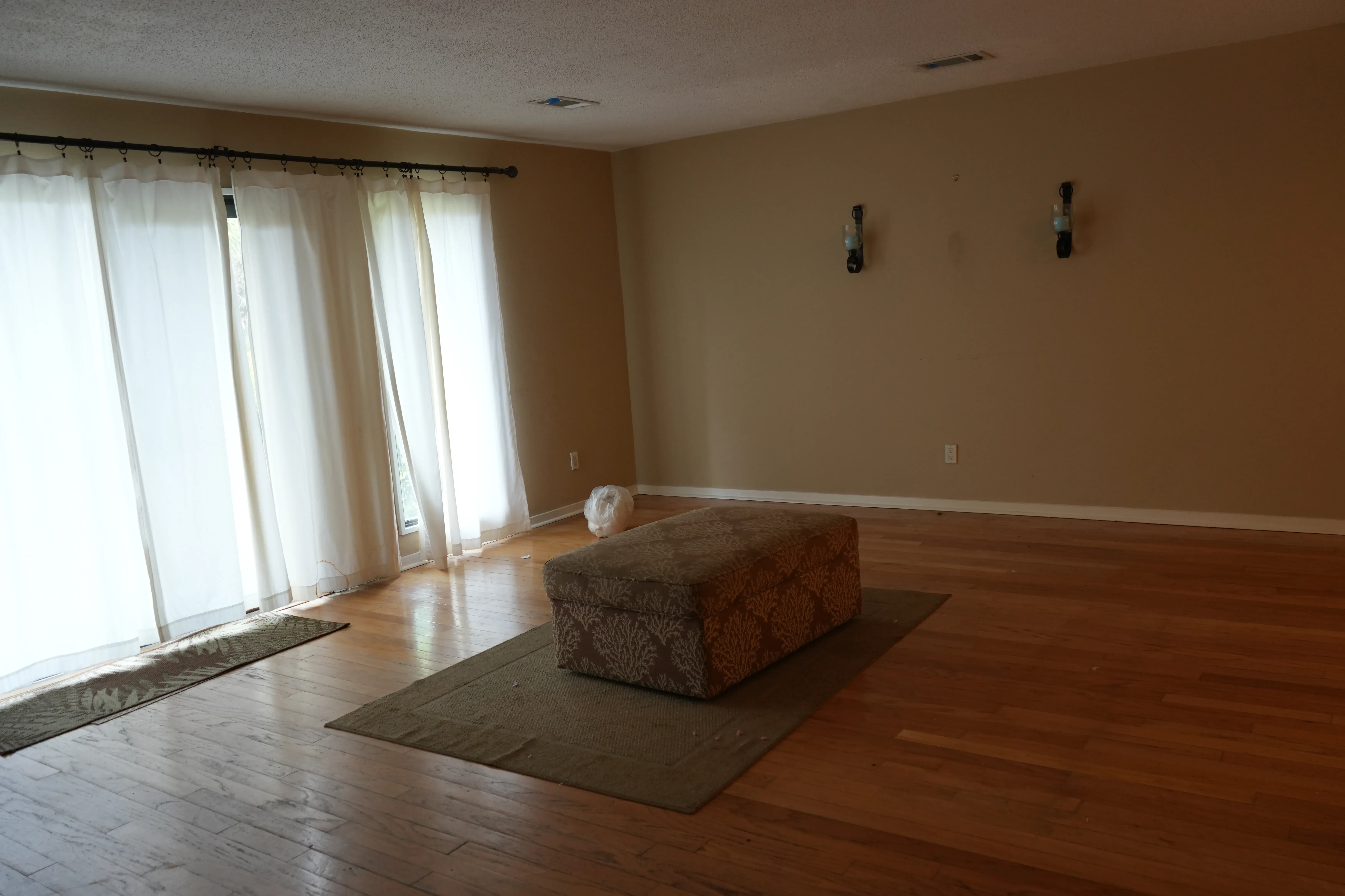 An empty living room with hardwood floors, sheer curtains over a sliding glass door, tan walls, and a single upholstered ottoman on a small rug before renovation.