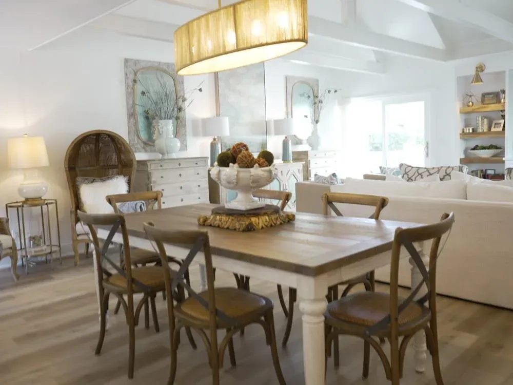 An open-concept dining area features a wooden farmhouse table with cross-back chairs beneath a large drum pendant light, with a vaulted beamed ceiling and a living room visible beyond.