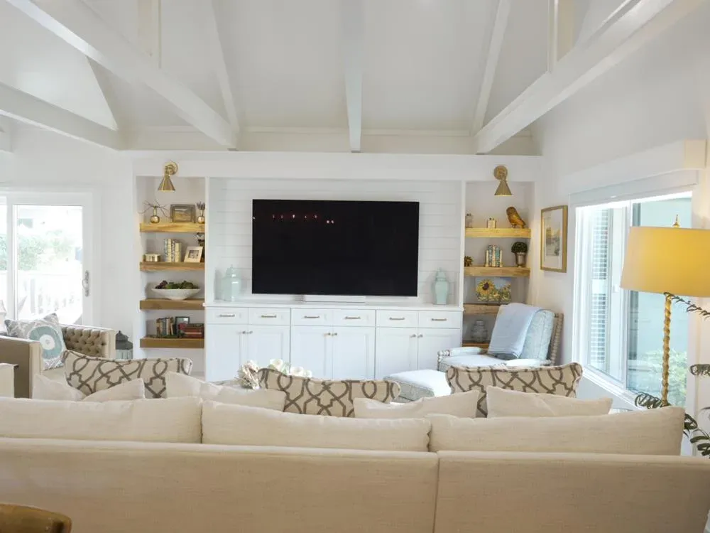 A bright living room with a vaulted white beamed ceiling features a built-in white cabinet flanked by open wood shelving and a mounted television, with a beige sectional sofa in the foreground and large windows letting in natural light.