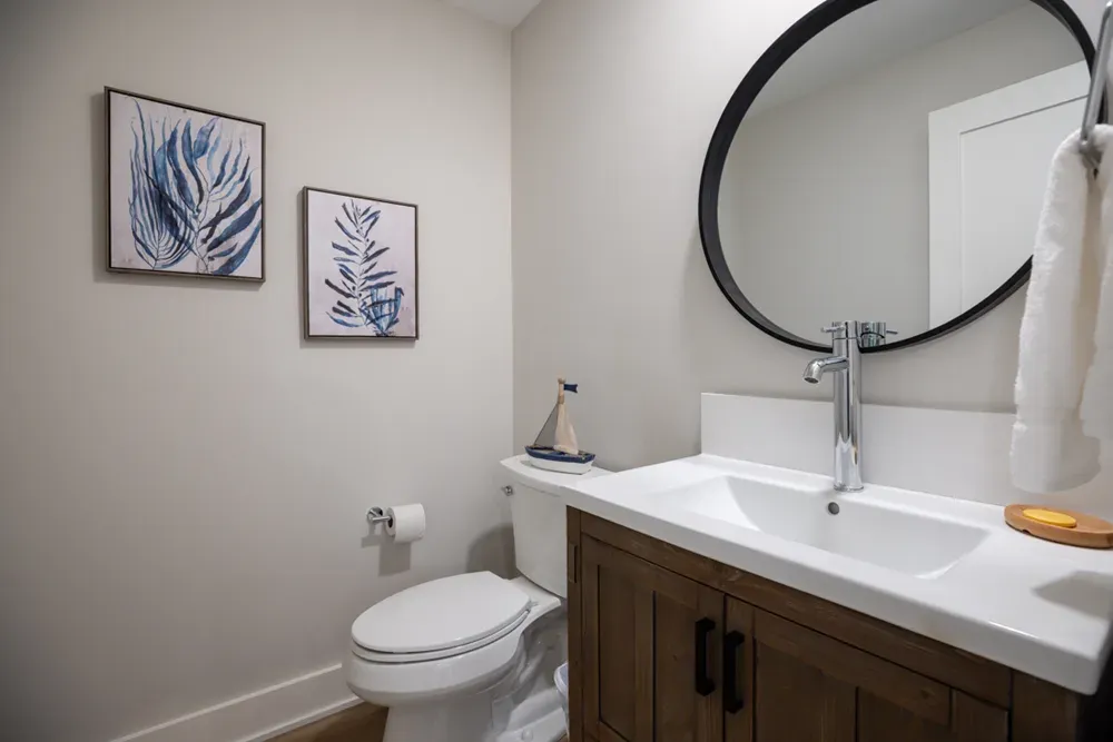 A small powder room features a wood vanity with a white countertop sink, an oval black-framed mirror, a toilet, and two framed botanical prints on the wall.
