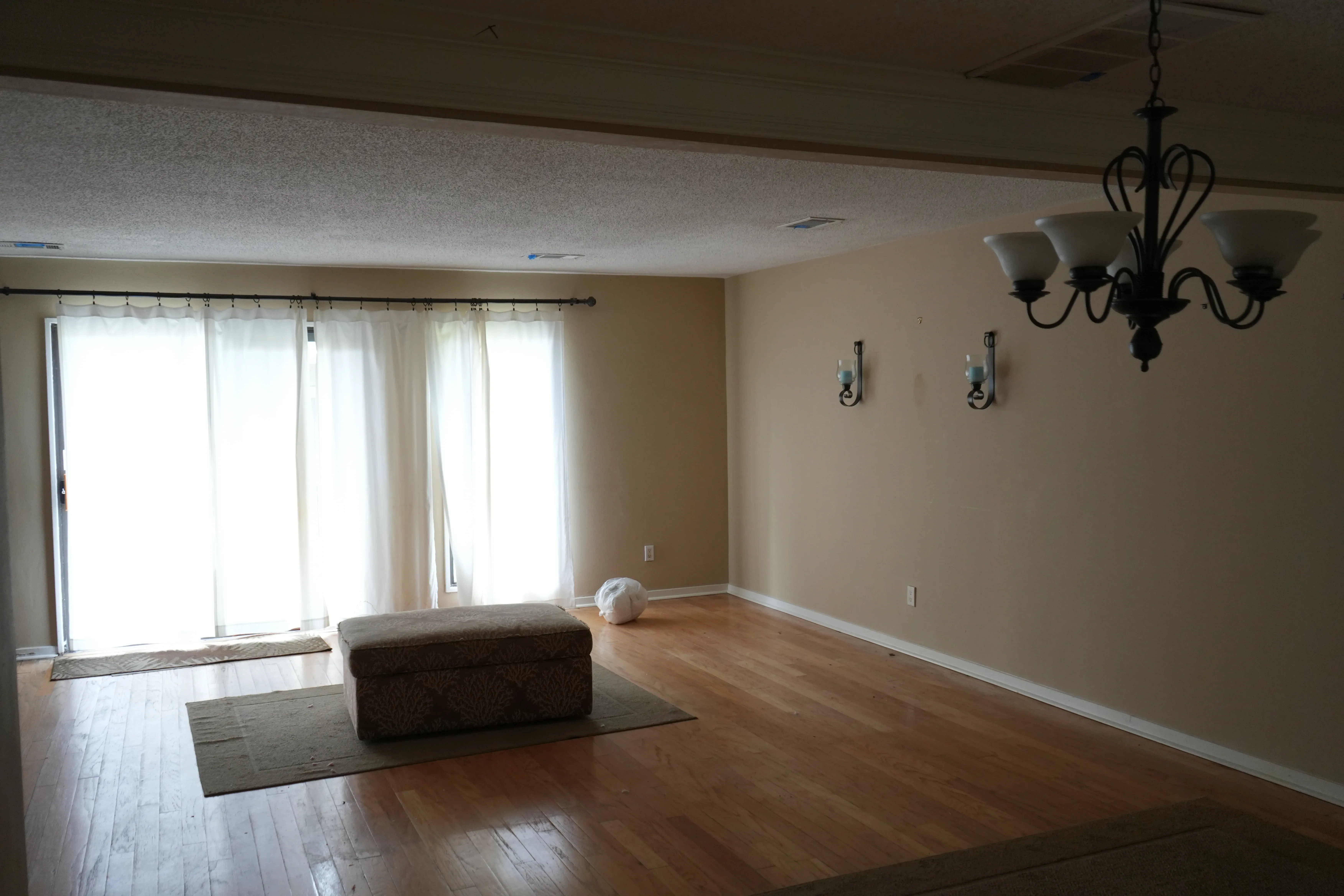An empty living and dining area before renovation with wood flooring, sheer curtains over sliding glass doors, wall sconces, and a wrought-iron chandelier hanging from a textured ceiling.