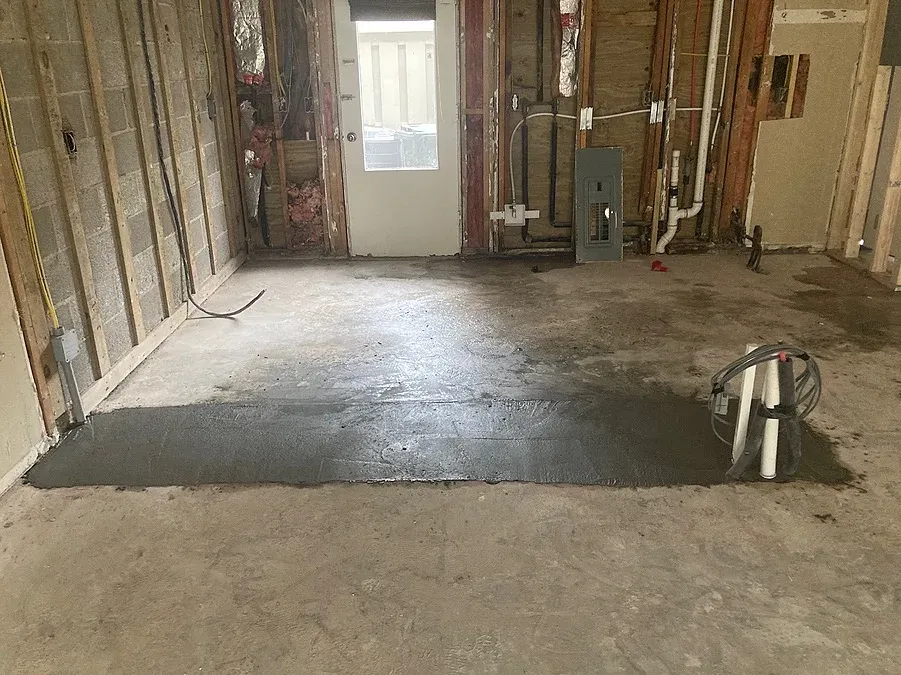 An under-construction room addition shows exposed wall framing, electrical panel, and plumbing rough-in around a freshly poured section of concrete floor.