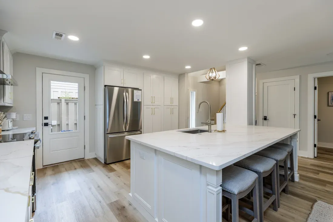 A remodeled kitchen features a large white marble-topped island with a sink and gray upholstered barstools, flanked by white shaker cabinets and a stainless steel refrigerator, with wood-look flooring extending toward a side door.