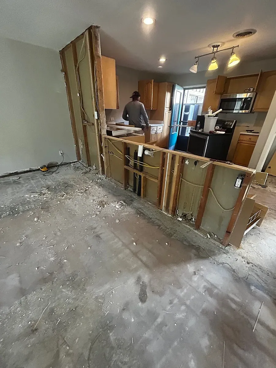 A kitchen mid-renovation shows exposed wall studs, a bare concrete subfloor covered in debris, and a worker standing near the original wood cabinets and appliances still in place.
