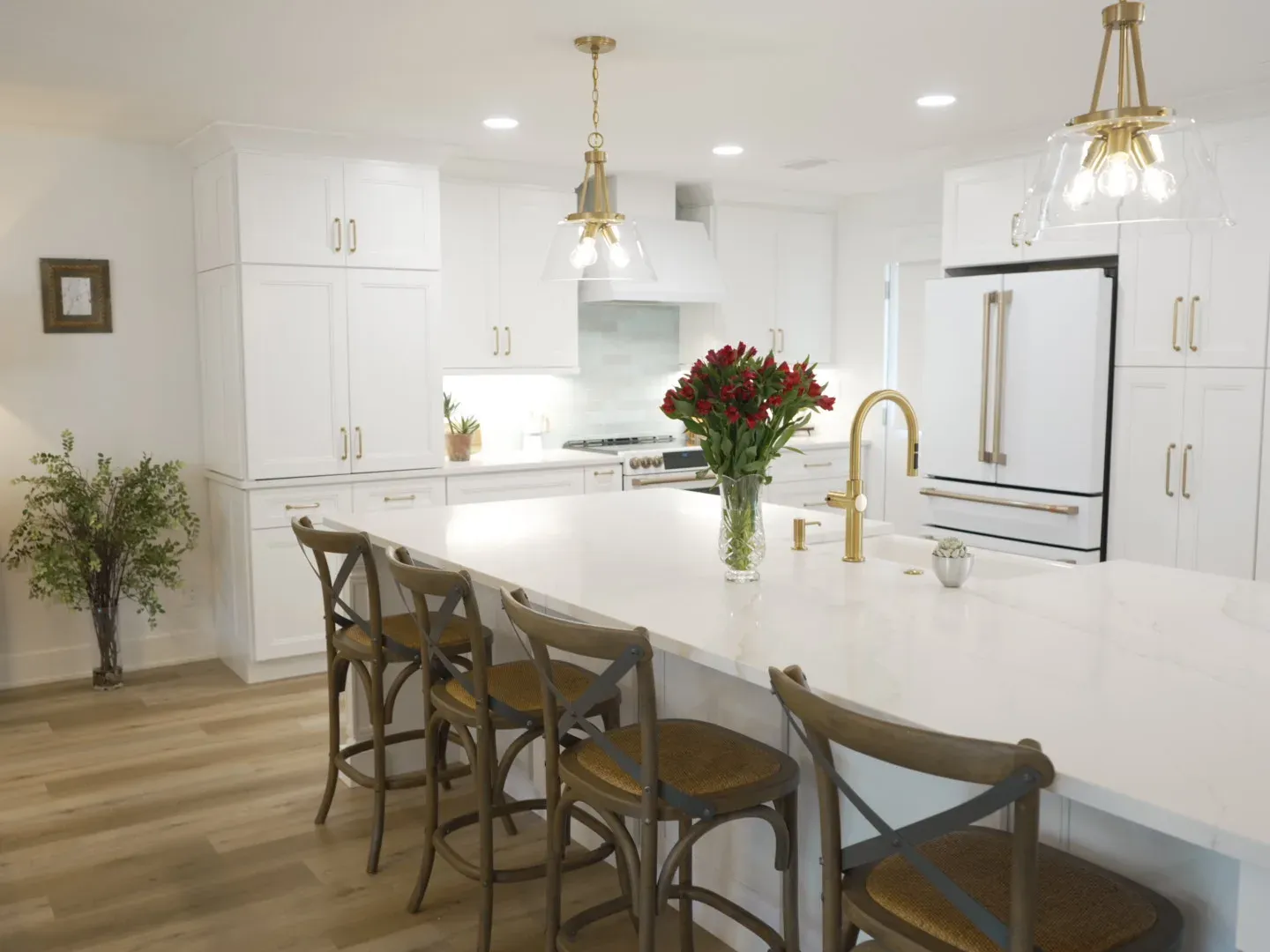 A bright white kitchen features a large quartz island with gold fixtures, cross-back barstools, and glass-shaded brass pendant lights above.