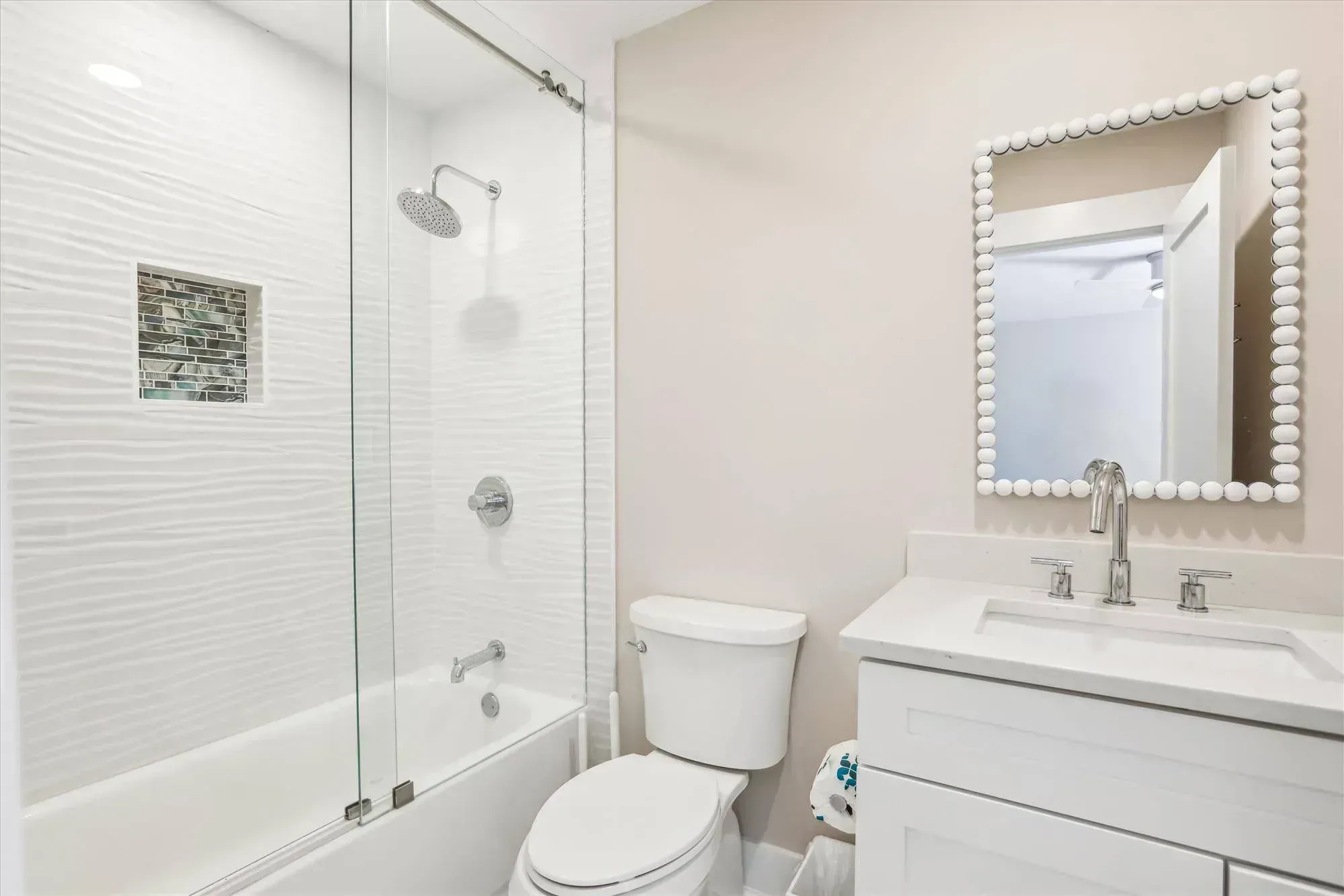 A remodeled bathroom features a tub and shower combination with textured white tile and a glass mosaic accent niche, alongside a white vanity topped with a beaded mirror and toilet.