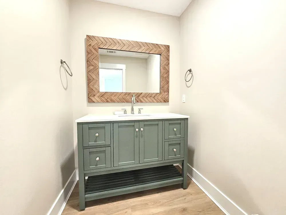A bathroom features a sage green vanity with a white countertop, brushed nickel fixtures, and a herringbone wood-framed mirror above a single undermount sink.