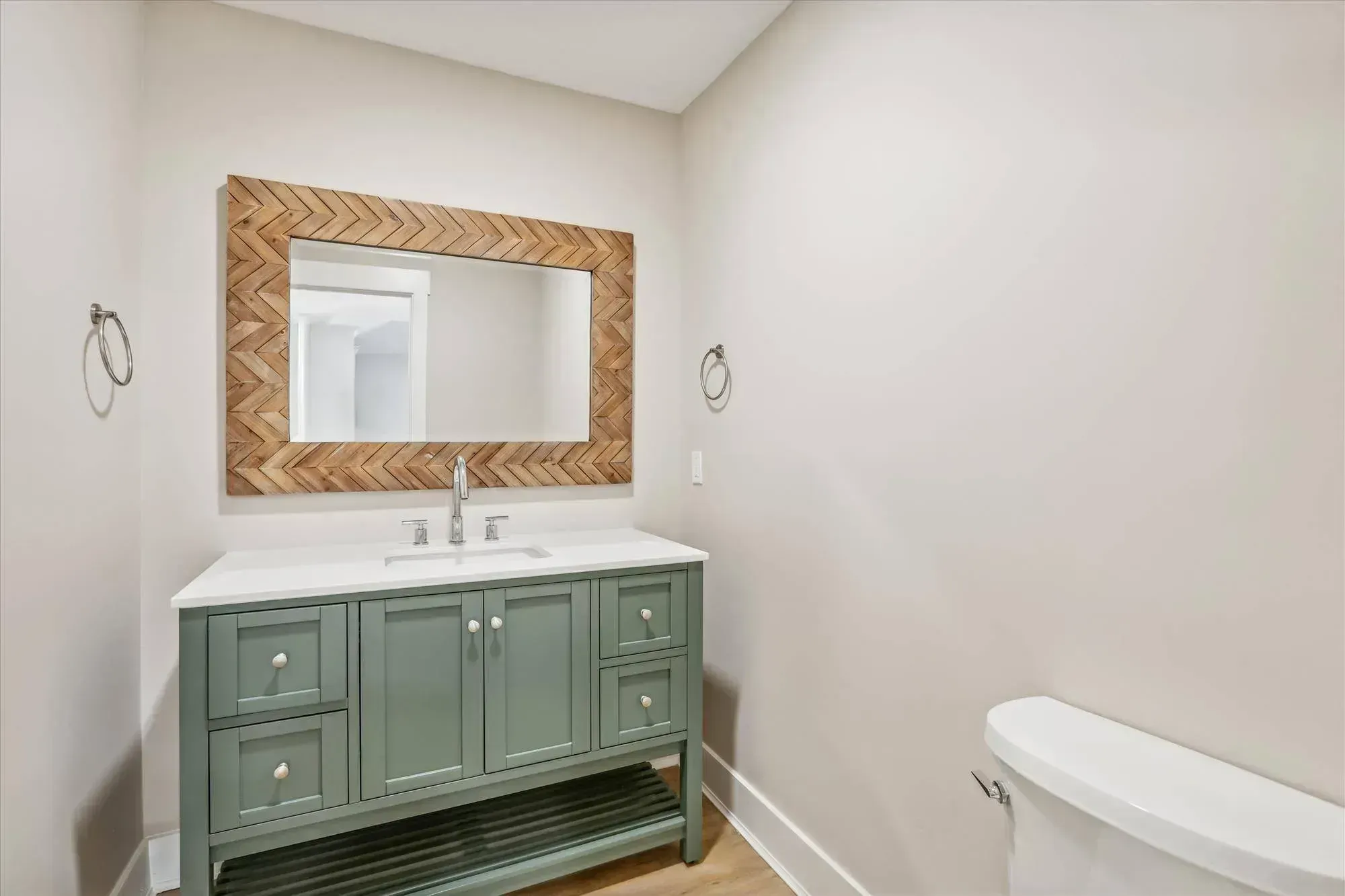 A bathroom features a sage green vanity with a white countertop and undermount sink beneath a large mirror framed in a herringbone wood pattern.