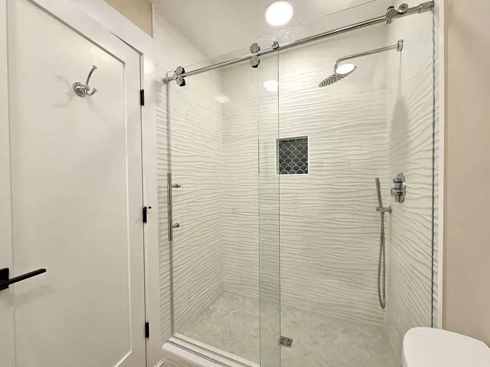A frameless glass shower enclosure with textured white wave-patterned tile walls, a rainfall showerhead, a handheld sprayer, and a small scalloped-tile niche above a herringbone tile floor.