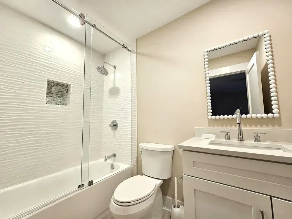 A remodeled bathroom features a tiled tub and shower enclosed by a frameless glass sliding door, alongside a white vanity with a beaded mirror above it.