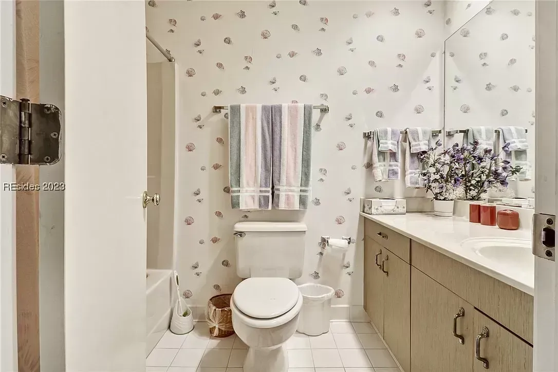 A bathroom with shell-patterned wallpaper features a wood-tone vanity with white countertop, a toilet, and a tub-shower combination visible through the open door.
