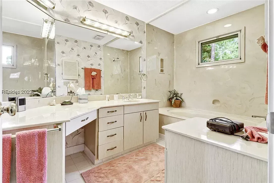 An outdated bathroom features a large mirrored vanity with floral wallpaper, light wood cabinetry, a built-in soaking tub beneath a window, and a pink shag rug on tile flooring.