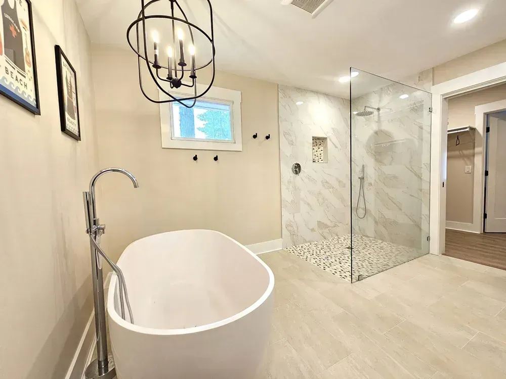 A remodeled bathroom features a freestanding oval soaking tub with a floor-mounted faucet beside a walk-in glass shower clad in marble-look tile, all lit by a black candelabra-style chandelier.